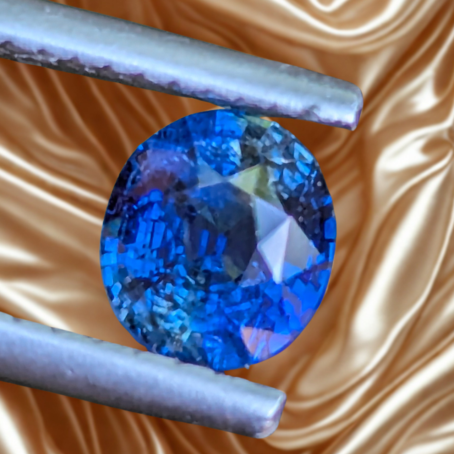 A oval-cut blue gemstone held in silver-colored metal tweezers against a shiny gold satin fabric background.